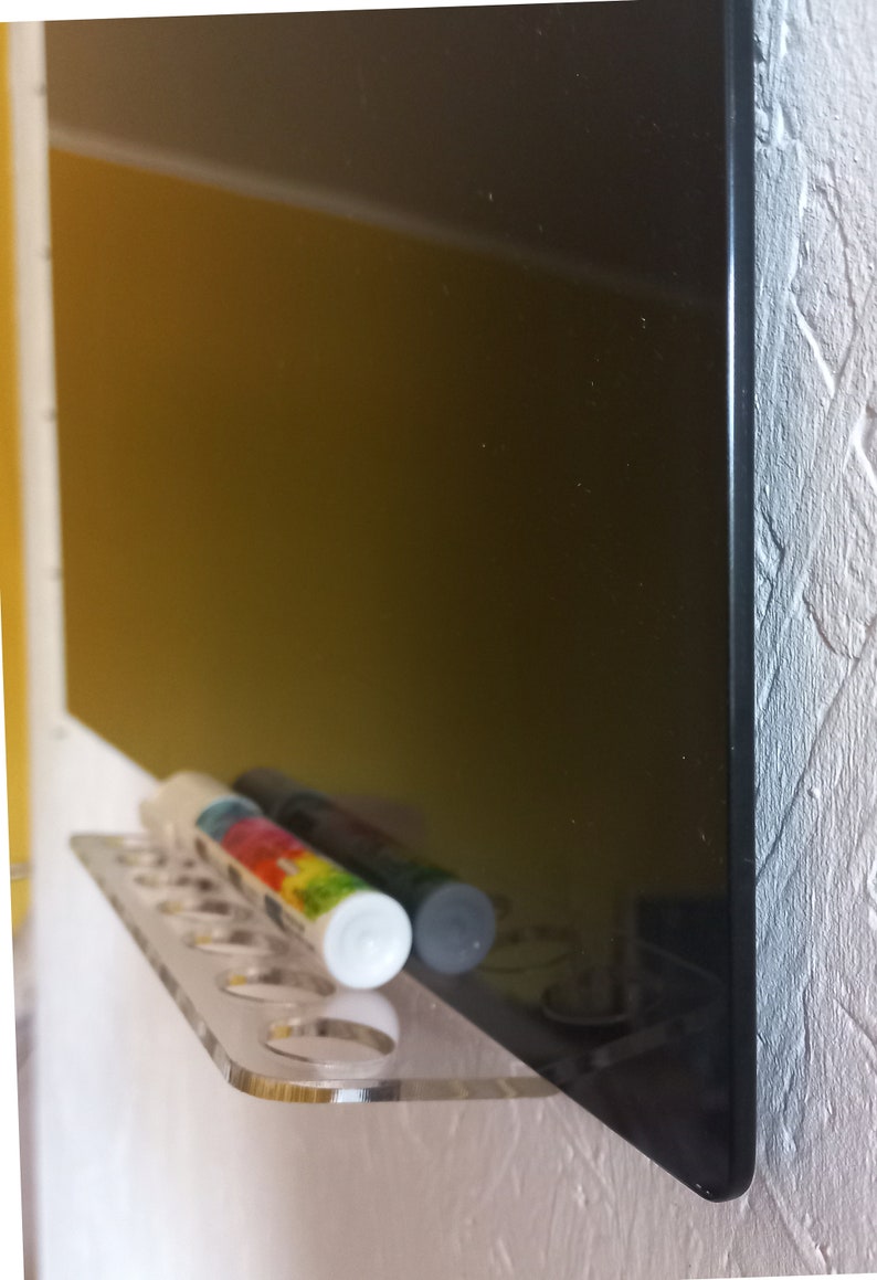 May include: A clear acrylic shelf with several round holes, holding two cylindrical markers. One marker has a colorful label, the other is dark. A large black surface is behind the shelf, reflecting the markers. The shelf is mounted on a white wall.