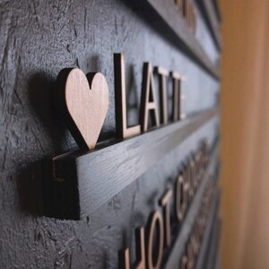 May include: A black wooden sign with the word "LATTE" in gold letters and a small wooden heart. The sign is mounted on a wall with other signs that say "HOT CHOCOLATE" and "APPLE PIE".