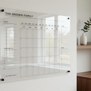 May include: A clear acrylic wall calendar featuring black printed text and grid lines. The header reads THE BROWN FAMILY. It includes sections for notes, a monthly calendar grid, a to-do list, a shopping list, and a menu, mounted with black hardware.