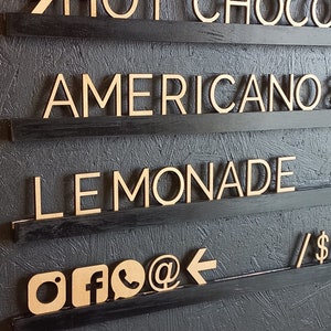May include: A black wooden sign with four shelves. The shelves have wooden letters spelling out "HOT CHOCOLATE", "AMERICANO", "LEMONADE", and social media icons.