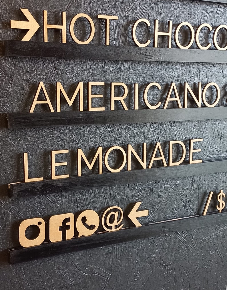 May include: A black wooden sign with four shelves. The shelves have wooden letters spelling out "HOT CHOCOLATE", "AMERICANO", "LEMONADE", and social media icons.