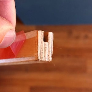 May include: Close-up of a wooden piece with a U-shaped cutout. The wood is light brown and has a natural grain. A piece of red tape is visible on the top of the wood.