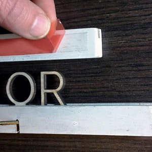 May include: Close-up of the word "OR" made of wood letters. The letters are attached to a white surface with a red adhesive strip.