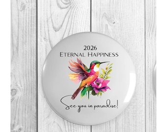 May include: A round white button featuring a colorful watercolor hummingbird hovering near a pink flower. The text 2026 ETERNAL HAPPINESS is printed above the bird, and the text See you in paradise! is printed in a cursive script below the bird.
