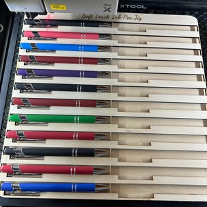 Puede incluir: Una colección de bolígrafos de colores en una plantilla de madera. Los bolígrafos tienen detalles plateados y están disponibles en varios colores, como negro, rojo, azul y verde. El texto "Soft Touch Ink Pen Jig" es visible.