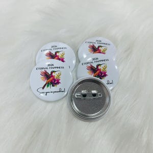 Puede incluir: Cuatro chapas redondas blancas con una ilustraci&oacute;n de un colibr&iacute; colorido, flores rosas y el texto 2026 ETERNAL HAPPINESS y See you in paradise! Una chapa est&aacute; dada la vuelta para mostrar la parte trasera de metal plateado con un alfiler.