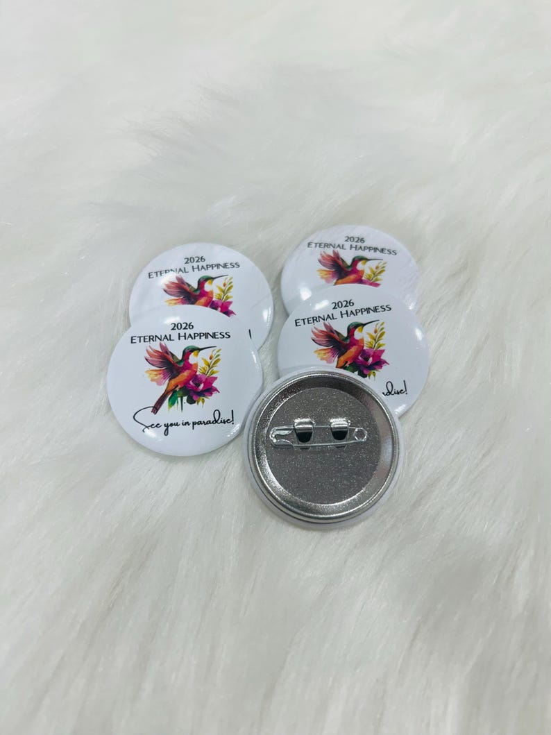 Puede incluir: Cuatro chapas redondas blancas con una ilustraci&oacute;n de un colibr&iacute; colorido, flores rosas y el texto 2026 ETERNAL HAPPINESS y See you in paradise! Una chapa est&aacute; dada la vuelta para mostrar la parte trasera de metal plateado con un alfiler.