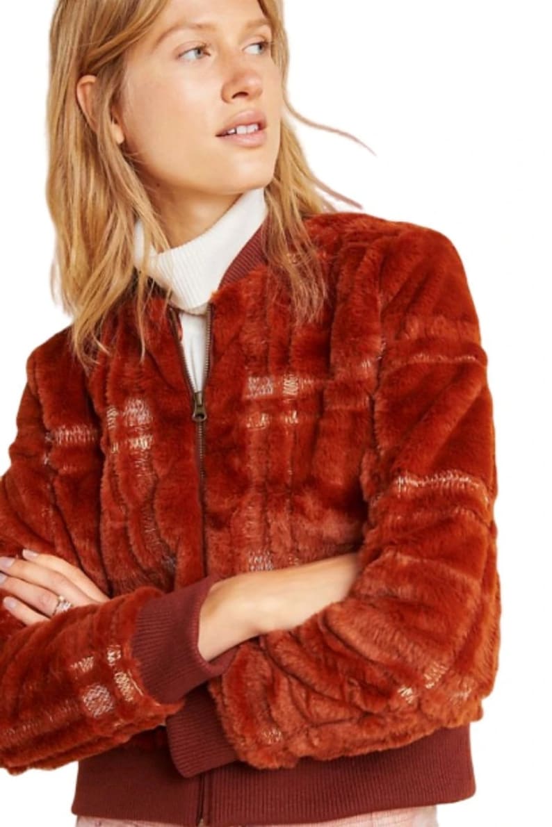 May include: A rust-colored faux fur bomber jacket with a plaid pattern. The jacket has a zipper closure and ribbed cuffs and hem.