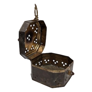 May include: An antique, octagonal metal box with a hinged lid. The box is open, revealing the interior. The lid and body feature decorative diamond-shaped cutouts. A small handle is attached to the top. The metal has a tarnished, aged appearance.