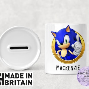 Könnte beinhalten: Weiße Keramik-Sparbüchse mit einem Schlitz oben. Die Sparbüchse hat ein blaues und goldenes Design mit einem Cartoon-Bild von Sonic the Hedgehog. Der Text "Mackenzie" ist auf der Vorderseite der Sparbüchse aufgedruckt.