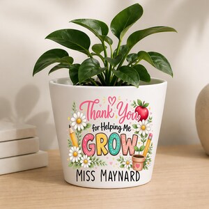 Può includere: Un vaso da fiori in ceramica bianca contenente una pianta verde. Il vaso presenta una grafica colorata con il testo Thank You for Helping Me GROW e MISS MAYNARD, circondato da illustrazioni di margherite, una mela rossa e matite.