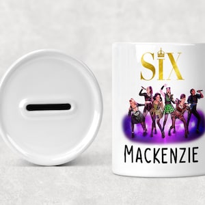Può includere: Una salvadanaio in ceramica bianca con una fessura, accanto a una banca cilindrica bianca con la parola "SIX" in oro, un gruppo di persone e il nome "Mackenzie" in nero. La banca ha uno sfondo viola e nero.
