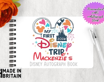 May include: A white spiral-bound autograph book featuring a colorful graphic of Mickey Mouse ears, a castle, and the text My First Disney Trip Mackenzie's Disney Autograph Book 2026. A clear pen is placed next to the book, which is A6 in size.