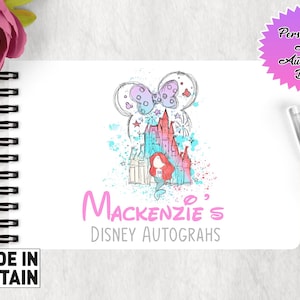 May include: A white A6 autograph book with a pink and blue watercolor illustration of Cinderella's castle and a pink bow. The text "Mackenzie's Disney Autographs" is printed in pink below the illustration.