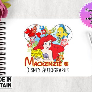 May include: A white A6 autograph book with a Disney theme. The cover features Ariel from The Little Mermaid, Sebastian the crab, and Flounder the fish. The text on the cover reads "Mackenzie's Disney Autographs".
