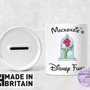 May include: White ceramic piggy bank with a slot on top. The front of the bank has a colorful image of a rose in a glass dome and the text "Mackenzie's Disney Fund".