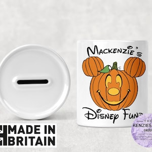 May include: A white ceramic money bank with a slot, next to a cylindrical bank decorated with a smiling pumpkin design. The text "Mackenzie's Disney Fund" is printed on the bank. The words "Made in Britain" are also visible.
