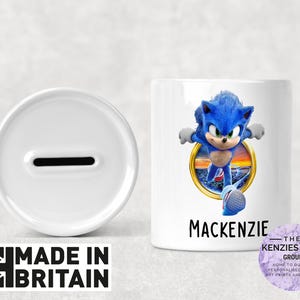 Könnte beinhalten: Weiße Keramik-Sparbüchse mit einem Schlitz oben. Die Sparbüchse hat ein blaues und weißes Bild von Sonic the Hedgehog auf der Vorderseite. Der Text "Mackenzie" ist unter dem Bild gedruckt.