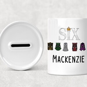 Könnte beinhalten: Weiße Keramik-Spardose mit einem Schlitz und eine passende zylindrische Bank mit dem Aufdruck "SIX" und "Mackenzie". Die Bank ist mit farbenfrohen Illustrationen von Kleidern verziert.