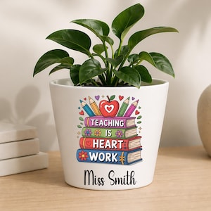 Puede incluir: Una maceta de cerámica blanca con una planta verde. La maceta presenta un gráfico colorido de una manzana, lápices y una pila de libros con el texto "TEACHING IS HEART WORK" y el nombre "Miss Smith" impreso en la parte inferior.