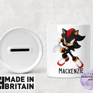 Può includere: Salvadanaio in ceramica bianca con uno slot nella parte superiore. La parte anteriore della scatola presenta un'immagine di cartone animato di Shadow the Hedgehog della serie di videogiochi Sonic the Hedgehog. Il testo "Mackenzie" è stampato sotto l'immagine.