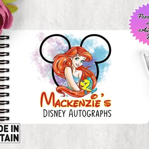 May include: A white A6 autograph book with a watercolor illustration of Ariel from The Little Mermaid holding Flounder. The illustration is surrounded by a Mickey Mouse ear shape. The text "Mackenzie's Disney Autographs" is printed below the illustration.