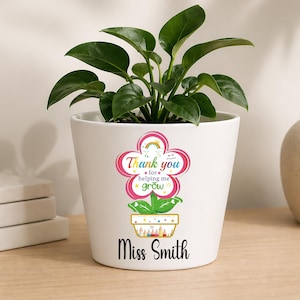 May include: A white ceramic flower pot with a green plant. The pot has a flower-shaped design with the text "Thank you for helping me grow" and "Miss Smith". The pot is on a wooden surface.