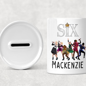 Può includere: Salvadanai in ceramica bianca con fessura e una banca cilindrica con la scritta "SIX" e una grafica a corona. Sotto, illustrazioni di sei donne in costumi colorati. Il nome "MACKENZIE" è stampato sotto.