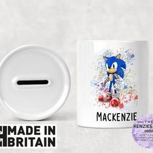 Könnte beinhalten: Weiße Keramik-Sparbüchse mit einem Schlitz oben. Die Vorderseite der Box zeigt ein farbenfrohes Bild von Sonic the Hedgehog mit dem Namen Mackenzie darunter. Der Text "The Kenzie's Card Group" ist auf einem violetten Hintergrund gedruckt.
