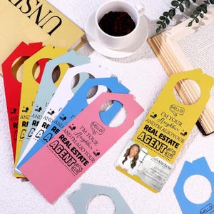 May include: A collection of house-shaped door hangers in red, yellow, light blue, white, and pink. Each hanger features the black printed text: Hello! I'm your neighbor and I'm also your real estate agent. One yellow hanger includes a business card.