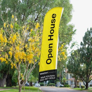 May include: A yellow and black open house sign with the text "Open House" in black lettering. The sign is attached to a pole and is standing in front of a tree with yellow flowers. The sign also includes the text "Welchert", "Matt Kitchens", and "562-567-SOLD".