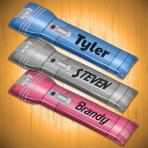 May include: Three flashlights in blue, silver, and pink, each featuring a black power button and a personalized name printed in black text. The blue flashlight is labeled Tyler, the silver one says STEVEN, and the pink one is marked Brandy.