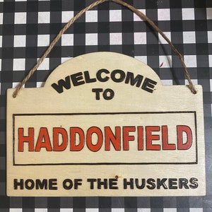 Könnte beinhalten: Ein Holzschild mit dem Text "Willkommen in Haddonfield, Heimat der Huskers" in schwarzer und roter Schrift. Das Schild hat einen Seilhalter.