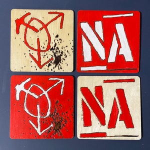 May include: Four wooden square tiles with red and white paint. Two tiles have the word "NA" in white letters on a red background. The other two tiles have a stylized symbol of a gender symbol with a red background and white symbol.