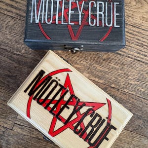 May include: Two wooden boxes with the band name "Motley Crue" and a red pentagram logo. The top box is dark gray wood and the bottom box is light wood.