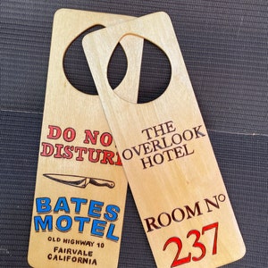 May include: Two wooden door hangers. The left one is for the Bates Motel, with the text "Bates Motel", "Old Highway 10", "Fairvale", and "California". The right one is for the Overlook Hotel, with the text "The Overlook Hotel", "Room No. 237".