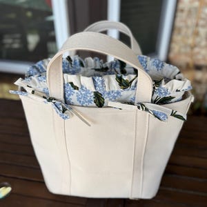 May include: Cream-colored tote bag with sturdy handles. The bag's interior features a floral pattern with blue flowers and green leaves. The top edge is ruffled, and the bag is tied with a matching floral ribbon. The bag is sitting on a wooden surface.