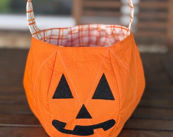 May include: A fabric pumpkin bucket featuring a bright orange exterior with a stitched jack-o'-lantern face made of black felt. The bucket has a rounded shape with vertical quilted lines and a handle lined with an orange and white plaid fabric.