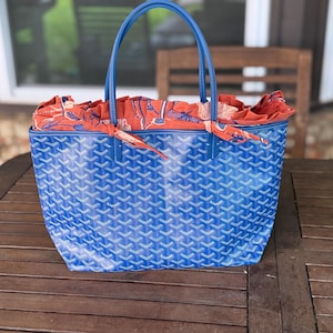 May include: A blue tote bag featuring a repeating geometric chevron pattern and the text Goyard Paris printed on the surface. The bag has two blue leather handles and an orange fabric insert with a floral pattern that extends from the top opening.