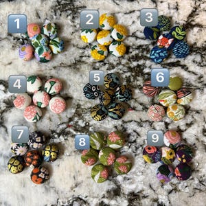 May include: Nine sets of fabric-covered buttons with floral patterns. The buttons are in various colors, including pink, yellow, blue, green, and orange. Each set has a different floral design, and the buttons are arranged in small clusters.