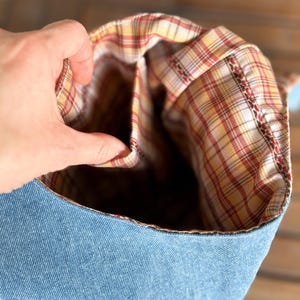 Puede incluir: Una bolsa de mezclilla con forro de tela a cuadros. El exterior de la bolsa es de mezclilla azul claro, y el forro interior presenta un patr&oacute;n de cuadros rojos, amarillos y blancos. La bolsa est&aacute; abierta, revelando el forro a cuadros.