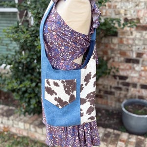 May include: A denim tote bag with a cowhide pattern panel and a brown pocket. The bag is paired with a floral print dress and top. The dress has a ruffled hem and the top has ruffled sleeves.