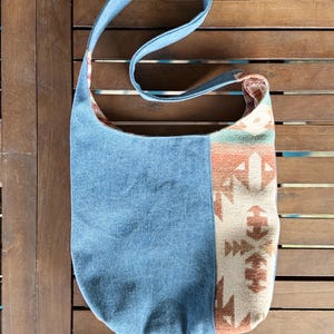 Puede incluir: Un bolso de hombro de mezclilla azul con un panel lateral estampado. El bolso tiene una correa larga y una forma rectangular. El panel estampado presenta dise&ntilde;os geom&eacute;tricos en tonos beige, naranja y verde.