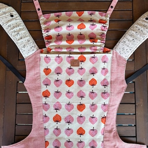 May include: A baby carrier with a strawberry pattern in pink and orange on a cream background. The carrier has black straps and a pink and white striped trim. The word "Happy" is on a small brown tag.