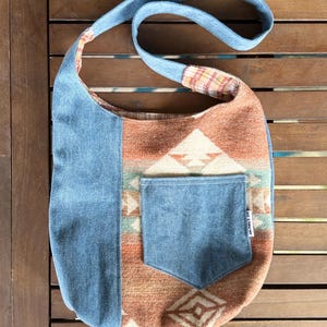 May include: A denim and patterned fabric shoulder bag. The bag features a light blue denim exterior with a pocket and strap. The main body of the bag has a geometric pattern in shades of brown, orange, and white. The interior lining is a plaid pattern.