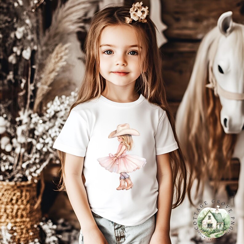 May include: A white short-sleeved t-shirt featuring a graphic illustration of a child wearing a cowboy hat, a pink tutu, and brown cowboy boots. The graphic is centered on the chest of the shirt, which is worn with light blue denim jeans.