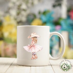 May include: A white ceramic mug featuring a printed illustration of a young person with long blonde hair wearing a light pink tutu dress and a tan cowboy hat. The person is also wearing tan cowboy boots. The mug has a standard handle on the right.