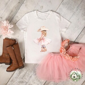 May include: A white short-sleeve t-shirt featuring a graphic of a girl wearing a cowboy hat, a pink dress, and boots. The set includes a pink tulle skirt, a floral headband, and a pair of brown leather-look boots with side zippers.