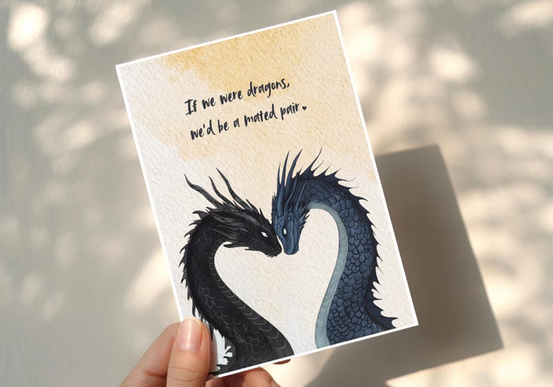 Op de afbeelding: Wenskaart met een drakenillustratie. Twee draken, &eacute;&eacute;n zwart en &eacute;&eacute;n blauw, vormen een hart. De tekst luidt: "Als we draken waren, zouden we een gepaard paar zijn."