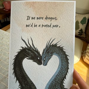 Op de afbeelding: Wenskaart met twee draken die een hart vormen. E&eacute;n draak is zwart, de andere blauwgrijs. De tekst luidt: 'Als we draken waren, zouden we een paar zijn.' De kaart heeft een achtergrond in aquarel-stijl.